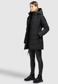 Kvinne iført en svart hette-quiltet vinterjakke, svarte leggings og svarte sneakers, stående med den ene hånden i lommene på jakken.