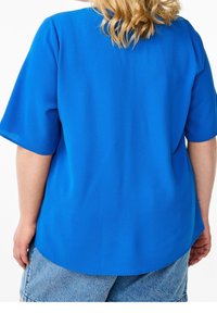 Sininen lyhythihainen paita, joka on valmistettu rakenteellisesta kankaasta. Paitassa on pyöreä kaula-aukko ja pyöristetty helma. Yhdistetty vaaleisiin farkku-shortseihin.