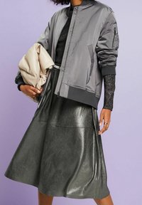 Veste bomber en satin gris avec poignets et taille côtelés, associée à une jupe évasée verte métallique ; tenant une pochette crème.
