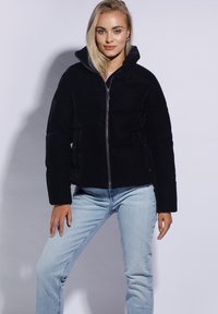 Sort, fyldt pufferjakke med høj krave og lynlås, der har sidelommer og en tætsiddende silhuet, parret med lyseblå jeans.