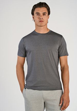 Camiseta gris de manga corta con cuello redondo, hecha de una tela texturizada. Combinada con pantalones de chándal gris claro, con las manos en los bolsillos.
