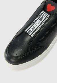 Zwarte leren sneaker met een gladde textuur, witte rubberen zool, geperforeerde neus en bedrukte labels met "LOVE MOSCHINO" en een rode hartaccent.