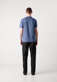 T-shirt de algodão azul com mangas curtas, combinada com calças de ganga escuras. A pessoa é vista de costas, destacando um design e um corte simples.