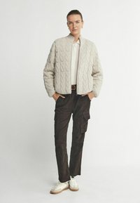 Ljus beige stickad bomberjacka med kabelmönster, krämfärgad blus, mörka cargo byxor och vita sneakers med bruna sulor.