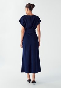 Morsko plava, duga haljina s kratkim rukavima i uskim strukom. Glatka tekstura, dužine do gležnjeva s detaljem zatvaranja na leđima. Jednostavan dizajn.