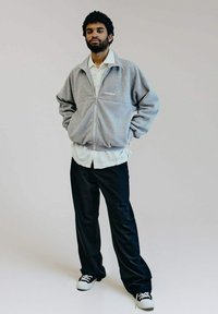 Graue Fleecejacke mit Reißverschluss und hohem Kragen, Logo auf der Brust, über einem weißen Shirt getragen, kombiniert mit lockeren schwarzen Hosen und Turnschuhen.