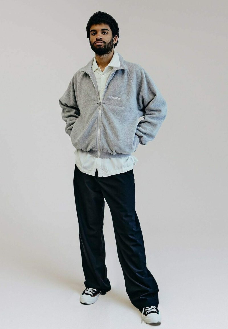 Graue Fleecejacke mit Reißverschluss und hohem Kragen, Logo auf der Brust, über einem weißen Shirt getragen, kombiniert mit lockeren schwarzen Hosen und Turnschuhen.