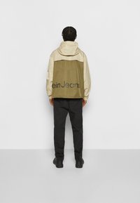 Beżowo-oliwkowa kurtka z kapturem z napisem "ein Jeans" na plecach, o luźnym kroju i lekkim materiale. Składana ze czarnymi spodniami.