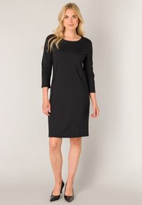 Zwarte knielange jurk met driekwart mouwen, ronde halslijn, gladde stof en een nauwsluitend silhouet. Gecombineerd met zwarte stilettohakken.