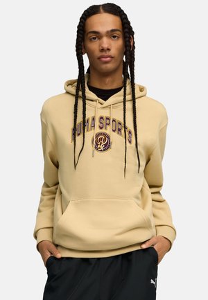 Giovane con trecce lunghe che indossa una felpa sportiva beige Puma e pantaloni neri Puma, mani in tasca, sfondo neutro.