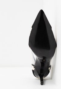 Chaussure à talon haut noire pointue avec panneaux latéraux transparents et accents à motif de cuir de vache, vue de la semelle sur fond blanc.