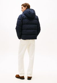 Uomo che indossa un giubbotto puffo blu navy con cappuccio, pantaloni bianchi e scarpe in suede marroni, in piedi di spalle su uno sfondo bianco.