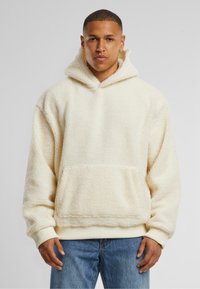 Cremfarbener, flauschiger Fleece-Hoodie mit großer Fronttasche, gerippten Bündchen und einer Kordelzugkapuze, getragen über blauen Jeans.