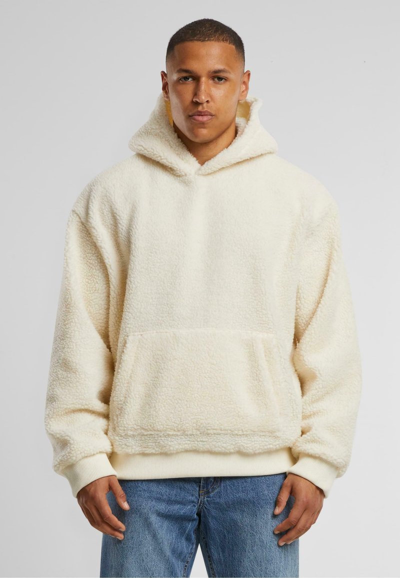 Cremfarbener, flauschiger Fleece-Hoodie mit großer Fronttasche, gerippten Bündchen und einer Kordelzugkapuze, getragen über blauen Jeans.