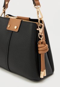 Czarna torebka o fakturze z wyraźną strukturą, z brązowymi akcentami, złotymi elementami i węzłowym detalem z boku. Zamek błyskawiczny na górze.