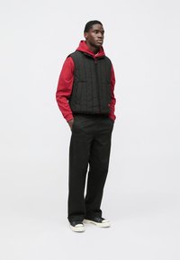 Gilet nero trapuntato sopra una felpa rossa con cappuccio, abbinato a pantaloni neri e sneakers nere con suola bianca. Design semplice con tasche pratiche.