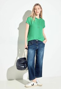 Felpa verde a maniche corte con cappuccio e cordino, jeans ampi blu, sneaker bianche con dettagli neri e una borsa a tracolla nera.