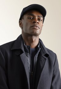 Abrigo de lana azul marino superpuesto sobre un suéter de punto azul marino con cremallera, que presenta un cuello y solapas al estilo de una chaqueta. Se lleva una gorra a juego.