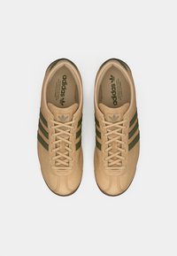 Ζευγάρι από μπεζ σουέντ αθλητικά παπούτσια Adidas με σκούρες πράσινες ρίγες και κορδόνια, σε θέα από πάνω σε λευκό φόντο.