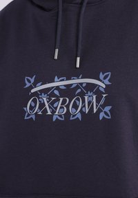 Hoodie marine avec un design graphique portant le mot « OXBOW » en argent, entouré d'accents floraux bleus et fermeture à cordon avec embouts en argent.