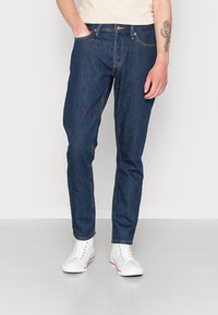 Jeans in denim blu scuro con taglio dritto, caratterizzati da cinque tasche, patta con bottone e dettagli leggermente consumati. Indossati con sneakers bianche.
