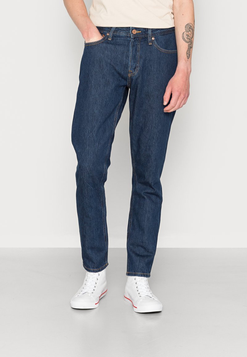 Jeans in denim blu scuro con taglio dritto, caratterizzati da cinque tasche, patta con bottone e dettagli leggermente consumati. Indossati con sneakers bianche.