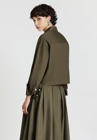 Camicia corta verde oliva con colletto, maniche lunghe e dettagli a bottoni, abbinata a una gonna plissettata coordinata. Texture liscia.