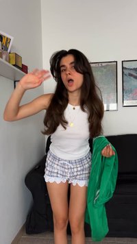 Weißes Tanktop, karierte Shorts mit Spitzenbesatz, hält eine grüne Jacke und steht in einem Raum mit einem schwarzen Sofa und gerahmter Kunst an der Wand.