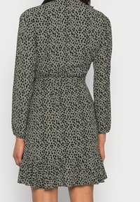 Green dress featuring a black spotted pattern, long sleeves, cinched waist, and a ruffled hemline. Made of a lightweight fabric.