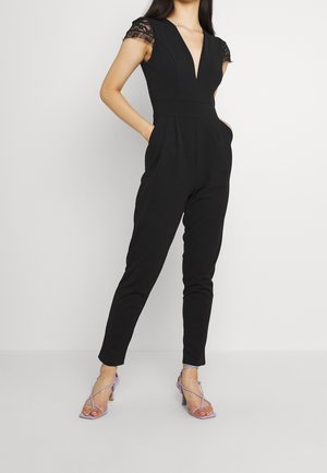 Jumpsuit noir avec un décolleté en V profond et des manches en dentelle. Il présente des jambes fuselées et des poches latérales. Associé à des escarpins à brides lavande.