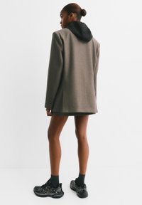 Blazer gris surdimensionné en tweed, avec une capuche noire. Le modèle le porte avec une jupe sombre et des baskets noires sportives.