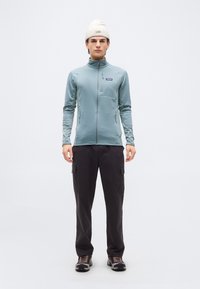 Νεαρός άνδρας στέκεται φορώντας ένα ανοιχτό γαλάζιο μπουφάν με φερμουάρ, μαύρο παντελόνι cargo, παπούτσια πεζοπορίας και λευκό πλεκτό σκουφάκι σε απλό φόντο.