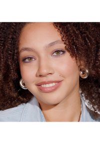 Porträtt av en person med lockigt hår, som bär silverfärgade hoops, lätt smink med lila detaljer och en denimjacka. Slät hy.