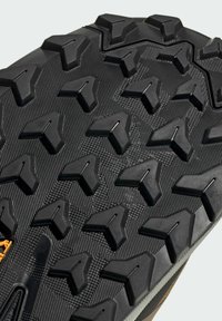 Black rubber outsole featuring pronounced triangular lugs and textured surface for traction. Reinforced edge with grey accents and orange logo.