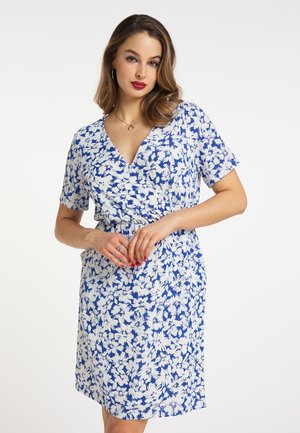 Vrouw draagt een blauw-wit bloemenprint korte mouwen wikkeljurk, poseert met lichtjes samengevouwen handen ter hoogte van de taille tegen een effen witte achtergrond.