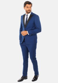 Completo da uomo blu con rever neri, design aderente e pantaloni slim. Abbinato a una camicia bianca e una cravatta blu scuro, scarpe nere lucide.