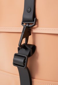 Une boucle noire attache une sangle texturée à un sac étanche de couleur pêche, présentant des surfaces lisses et continues ainsi qu'une couture contrastante.