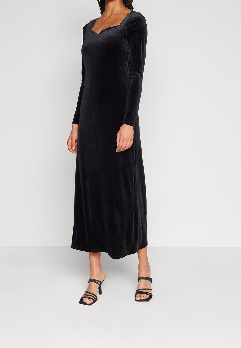 Robe longue en velours noir avec un décolleté carré, des manches longues et une coupe longueur sol. Portée avec des sandales noires à brides.