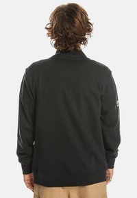 Black sweatshirt with long sleeves and a relaxed fit. Features a subtle collar and a label on the left sleeve. Soft, textured fabric.