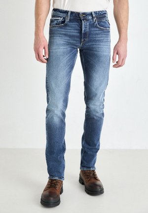 Uomo che indossa jeans blu skinny e stivali beige con lacci, in piedi su un pavimento chiaro uniforme davanti a uno sfondo bianco.