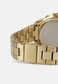 Zlaté kovové hodinky s designem náramku spojeného řetízkem. Obsahují sponu s vyrytým logem a hladké, odrazivé plochy. Minimalistické a odolné.