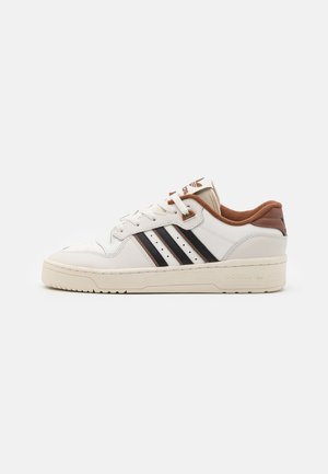 Baskets basses blanches à rayures noires, doublure marron, semelle beige et logo Adidas sur la languette et le talon, présentées en profil.