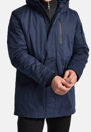 Man draagt een donkerblauwe winterjas met een ritszak op de borst en houdt zijn handen voor zich vast, zwarte broek zichtbaar eronder.