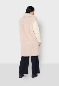 Faux fur vest in lichtroze, met een kraag en een vloeiende uitstraling, gelaagd over een crèmekleurige gewatteerde jas, in combinatie met zwarte wijde broekspijpen.