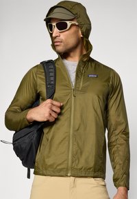 Man wearing olive green hooded jacket and cap, beige pants, white sunglasses, carrying black backpack over one shoulder.