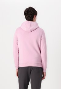 Hoodie rose clair avec une capuche à cordon de serrage, des bords côtelés et une texture lisse. Porté avec un pantalon gris, montrant une tenue décontractée.