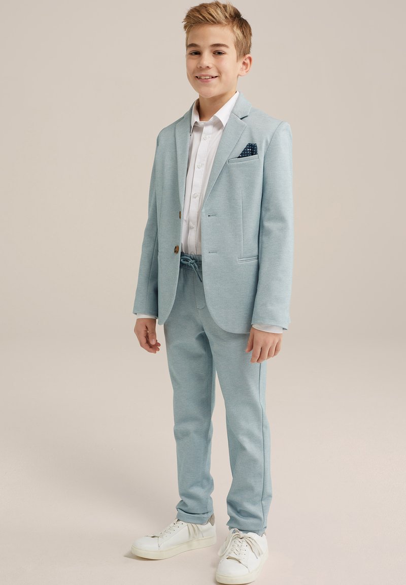 Svetlo modro obleko s krojenim sakoom in ujemajočimi hlačami, v kombinaciji z belim srajcem in belimi supergami. Žepni robček zagotavlja dodatno podrobnost.