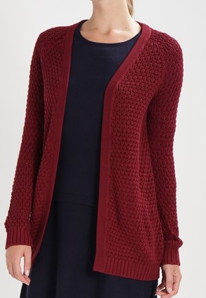 Kobieta w bordowym, teksturowanym kardiganie z długim rękawem, otwartym z przodu, na czarnej sukience na jednolitym tle.