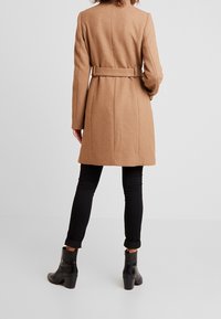 Kamelin värinen takki vyötärönauhan kanssa, muotoutunut malli, pitkät hihat ja takasauman yksityiskohdat, yhdistettynä mustiin kaarrettuihin housuihin ja nilkkasaappaisiin.