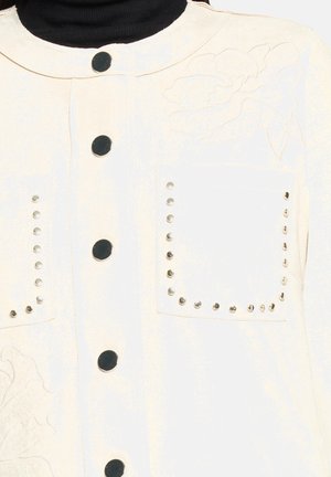 Veste blanche à col rond, avec deux poches avant ornées de clous et un motif floral embossé sur le côté gauche. Boutons noirs.